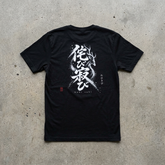 UTSURI WABI SABI (侘び寂び) t-shirt laid flat — the phrase wabi-sabi with a deer hidden in the brushstrokes across the back